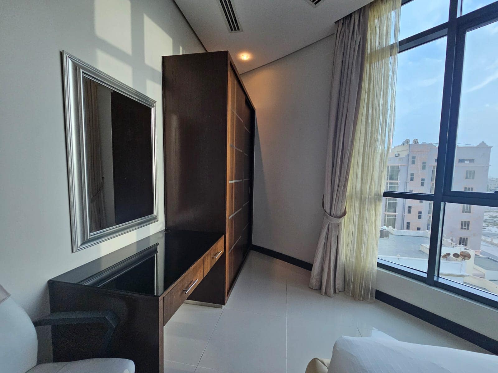 A sunlit corner of a modern room with a large window, long beige curtains, a wall-mounted mirror, a dark wooden cabinet, and a glass-topped desk.