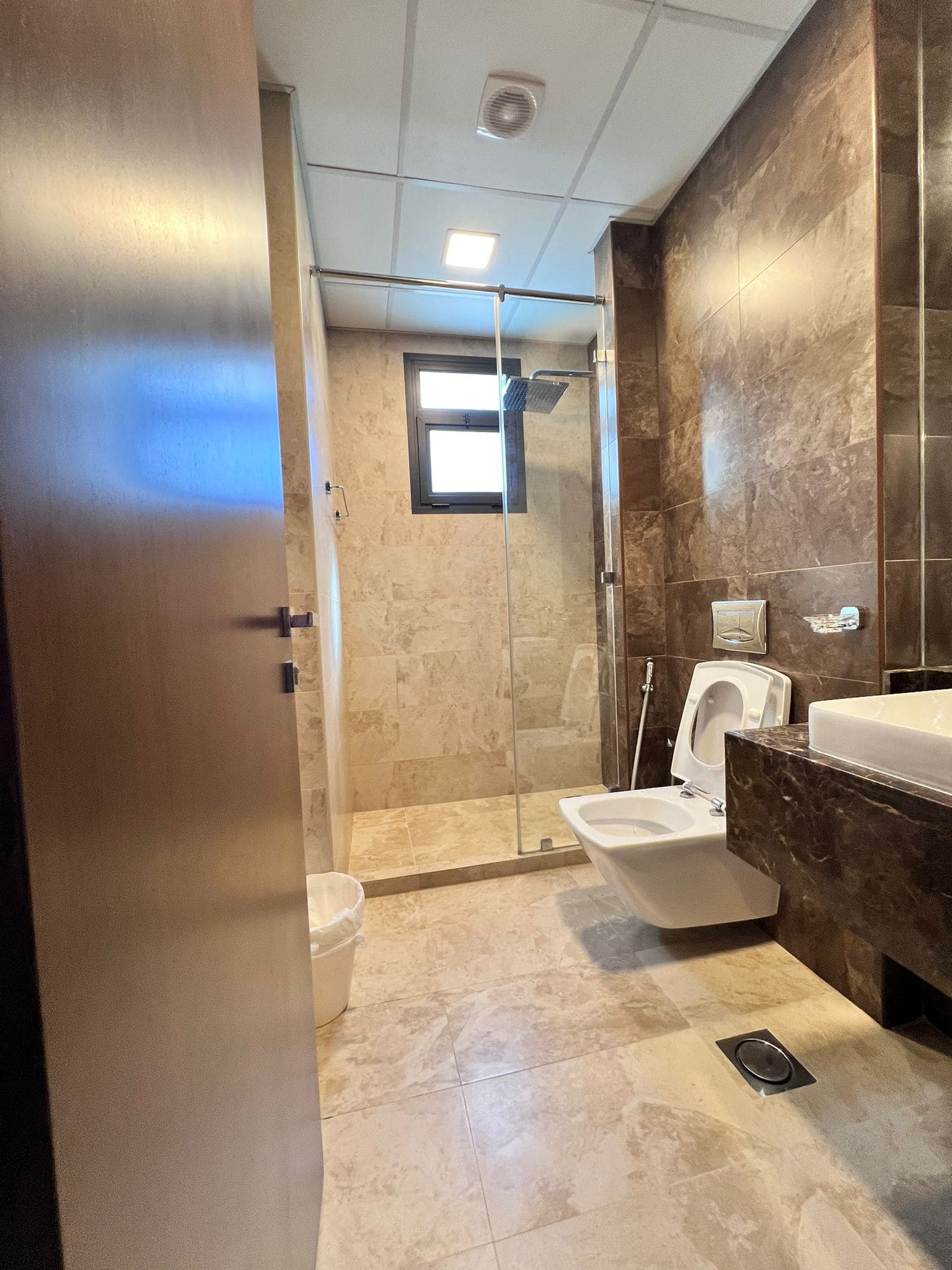 A modern bathroom in a Juffair flat for sale features beige marble tiles, a glass-enclosed shower, a wall-mounted toilet, a bidet, a sink with a counter, and a small trash bin.