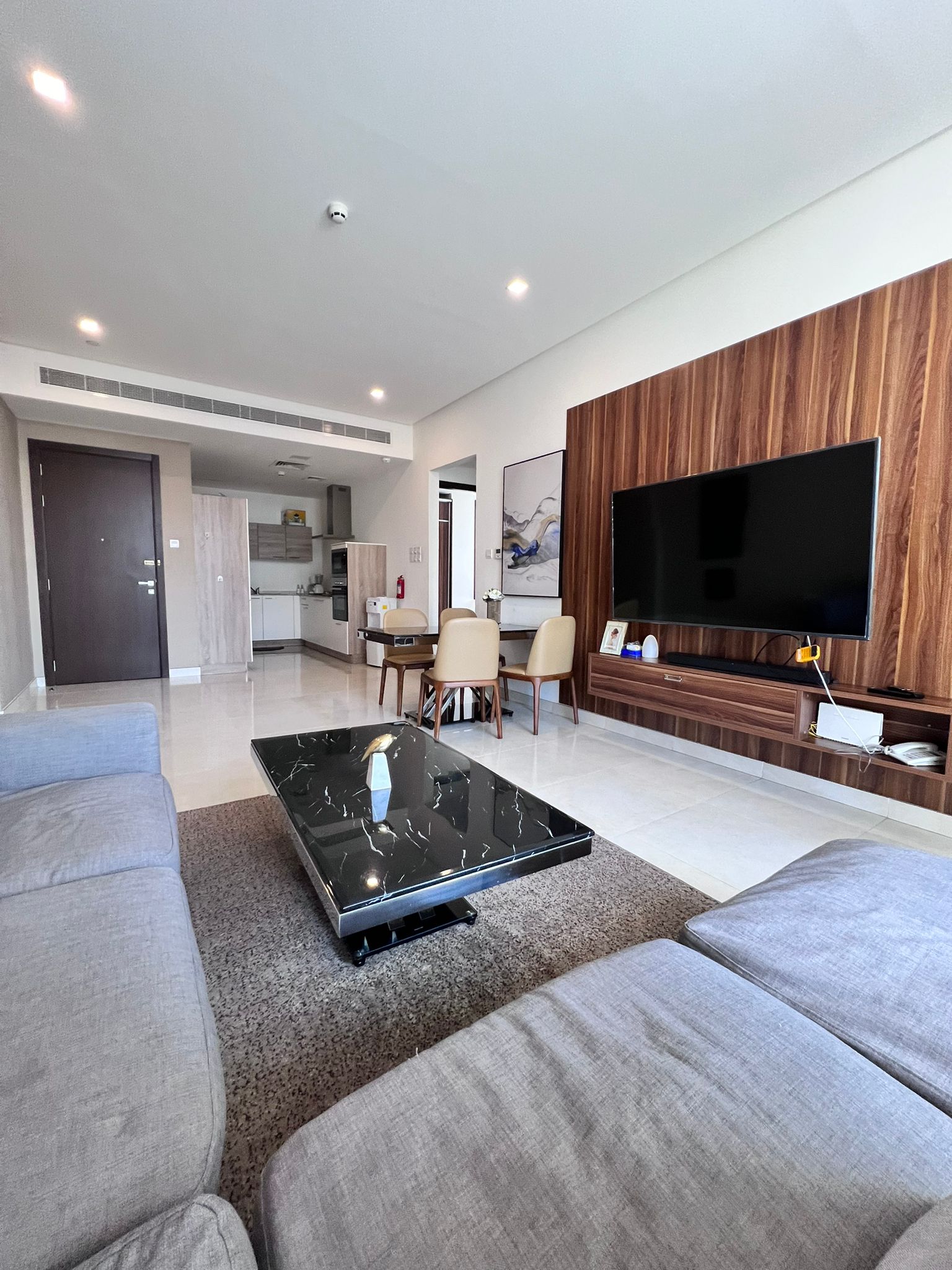 Modern living room in a flat for sale in Juffair, featuring a gray couch, black marble coffee table, and a large TV mounted on a wooden panel. The room opens to a dining area and kitchen, with neutral colors and minimalistic decor.