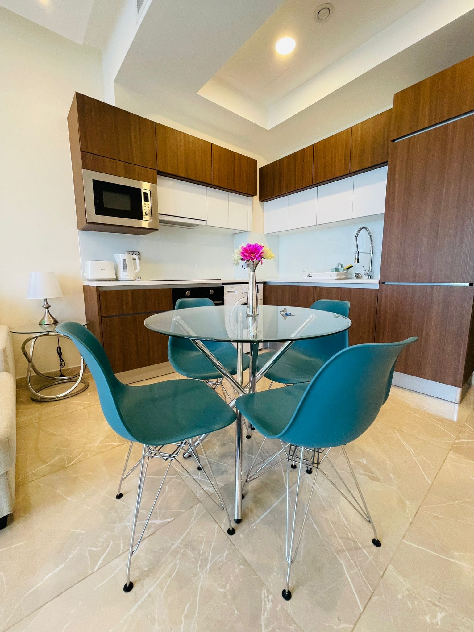 Apartment in Juffair: Modern kitchen with wooden cabinets, stainless steel appliances, and a glass dining table with teal chairs. A single pink flower in a vase adorns the table.