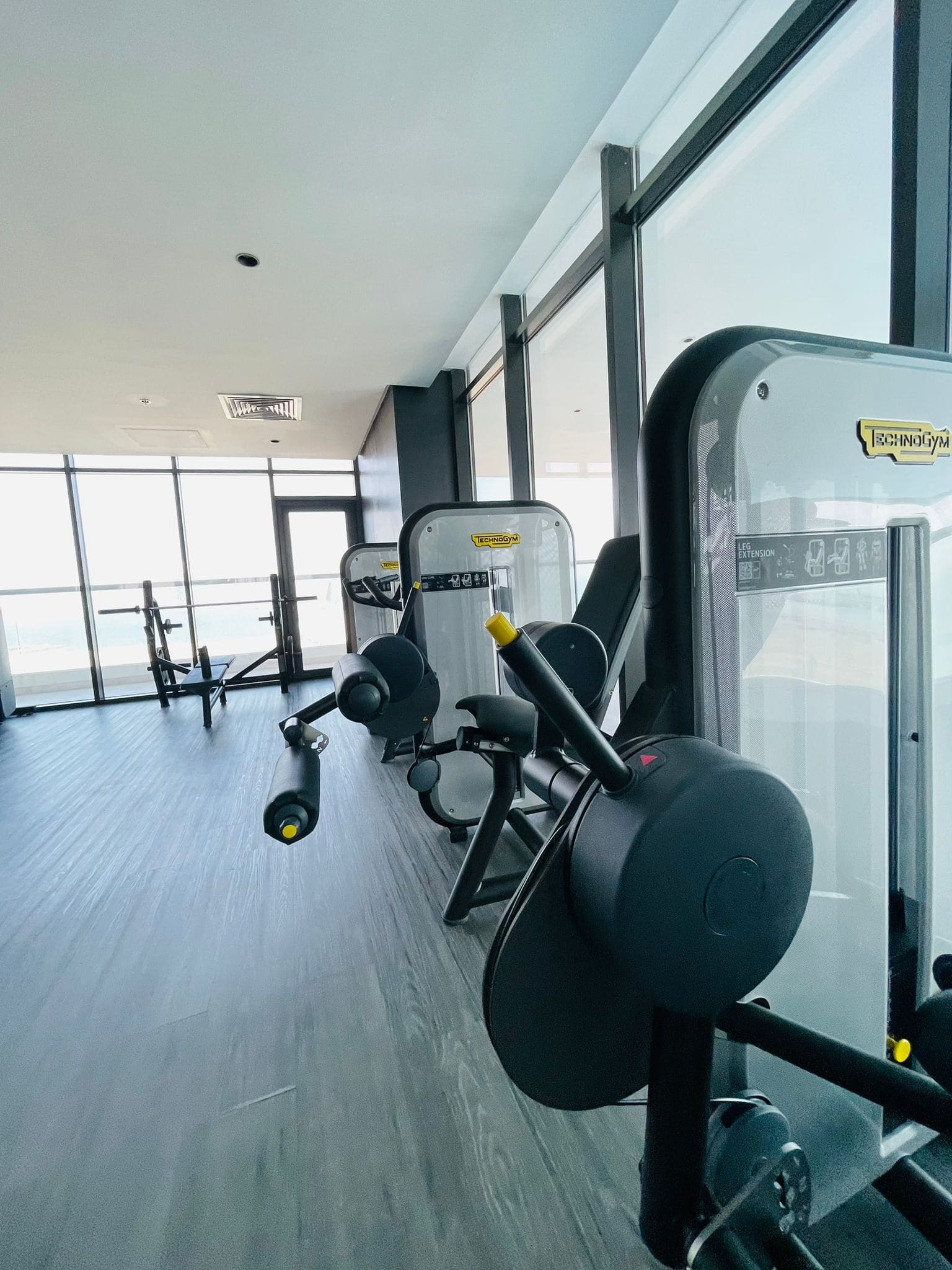 Modern gym interior within a luxury 1BR apartment in Juffair, featuring various workout machines including an elliptical, weight benches, and a weight machine, with large windows overlooking a city