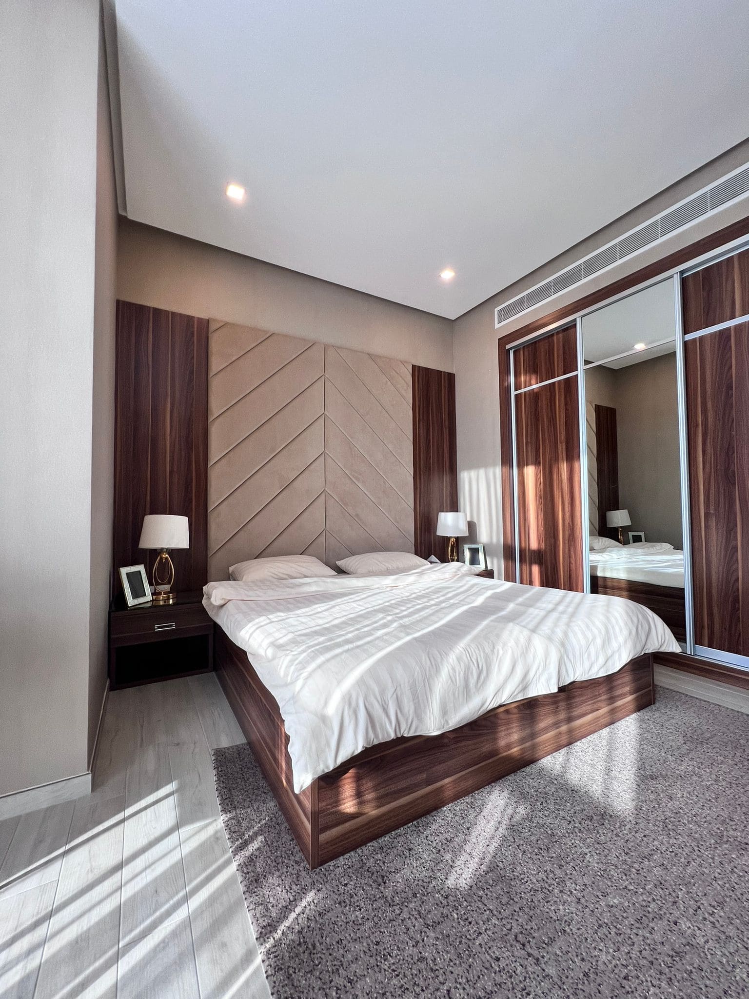 A modern bedroom in a flat for sale in Juffair, featuring a double bed with a wooden frame, two bedside tables with lamps and photos, a large mirror-front wardrobe, and gray carpeting.