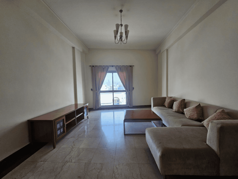 Flat for rent: Empty living room with a beige sectional sofa, a wooden coffee table, a tv console, and a chandelier, featuring large windows with a city view in Seqya.