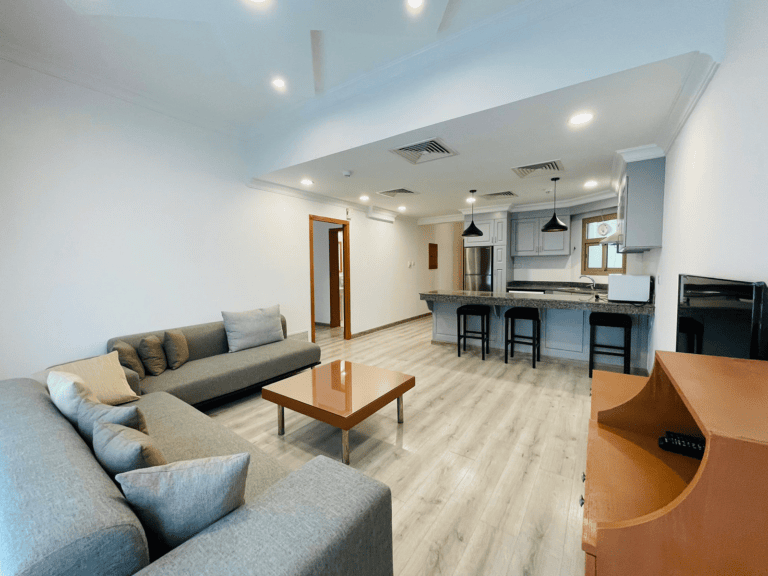 Modern living room with gray sectional sofa, wooden coffee table, open kitchen with bar, and bright lighting in a furnished apartment for rent in the Sanabis area.