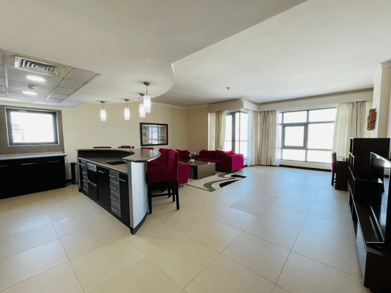 A modern, spacious luxury apartment living room with a kitchen counter, dining area, bright pink chairs, and large windows allowing natural light.