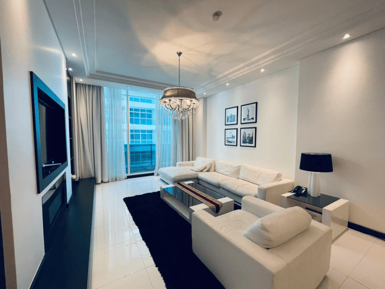 Luxury 2BR apartment living room interior with white sofas, a mounted tv, and a chandelier, with a city view through floor-to-ceiling windows.