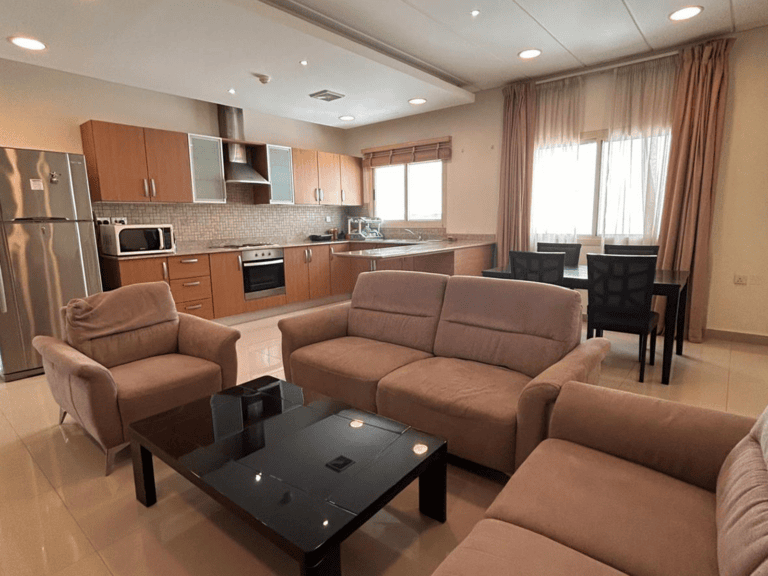 Modern living room connected to a kitchen with tan sofas, a glass coffee table, wooden cabinets, and stainless steel appliances in a 2 BDR apartment.