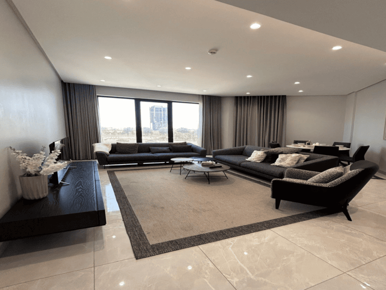 Modern living room in a luxury apartment with two dark sofas, a glass coffee table, and expansive windows offering a city view, complemented by neutral curtains and recessed lighting.