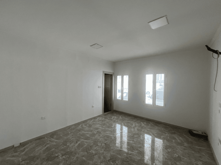Empty room with glossy tiled floor, white walls, a door, and two windows letting in natural light, available for rent in Al Hidd.