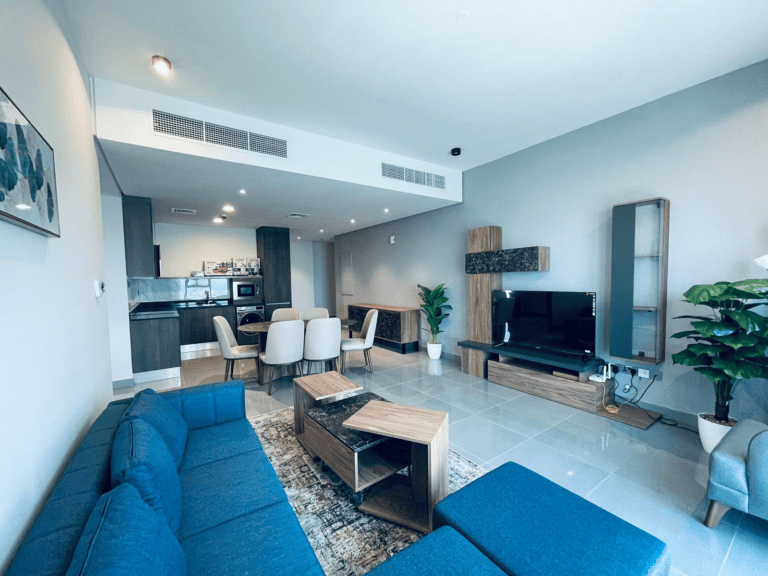 Modern apartment living room with blue sectional sofa, wooden coffee table, kitchen in background, and mounted tv.