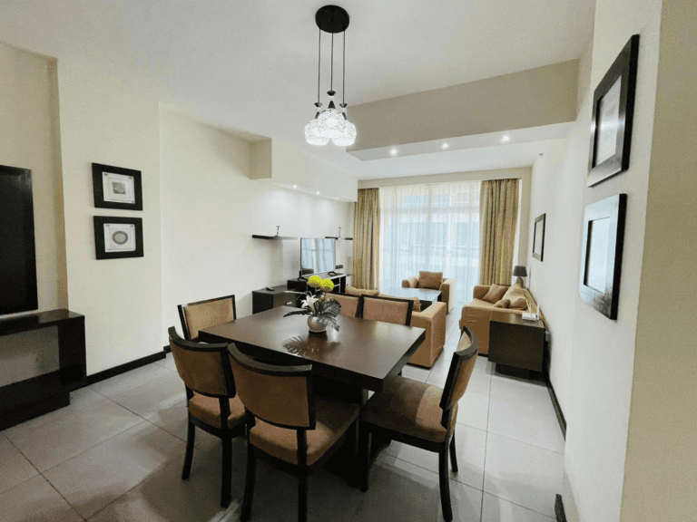 Luxury 2BR Apartment in Juffair with a modern dining and living room featuring a wooden table, brown chairs, a beige sofa set, and chandelier lighting.
