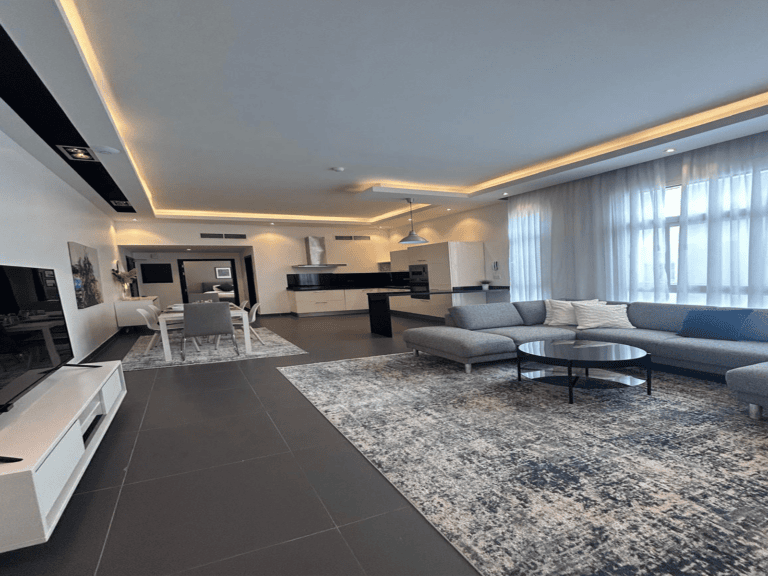 Modern living room with a large gray sectional sofa, an open kitchen layout, and ceiling accent lighting in a fully furnished luxury apartment.