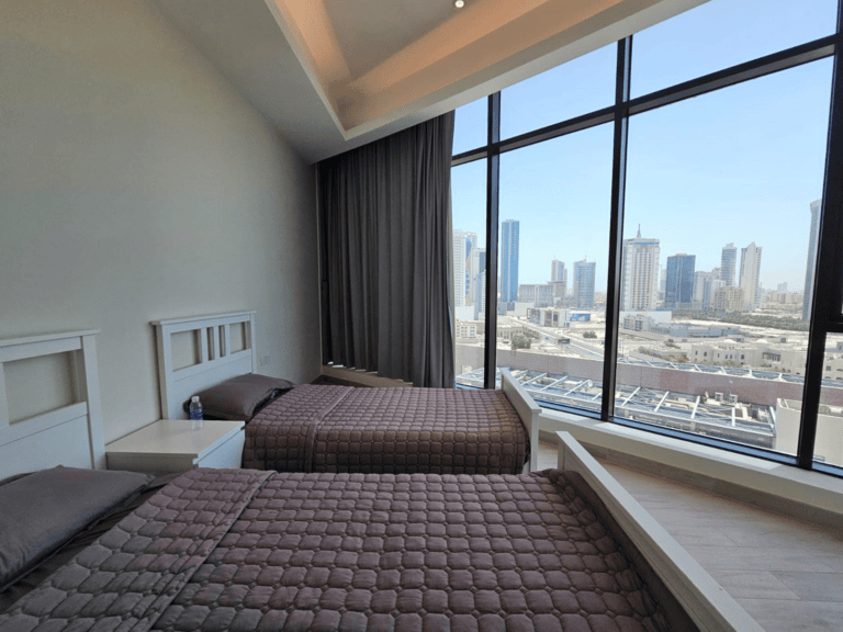 Modern three-bedroom apartment with a large window overlooking a city skyline, featuring a neatly made bed with brown bedding and elegant decor.