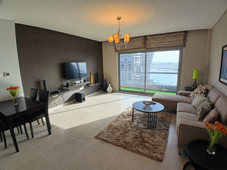 Modern living room in an Amwaj flat for rent, featuring a beige sofa, tv, and dining area, overlooking a view of the water through large windows.
