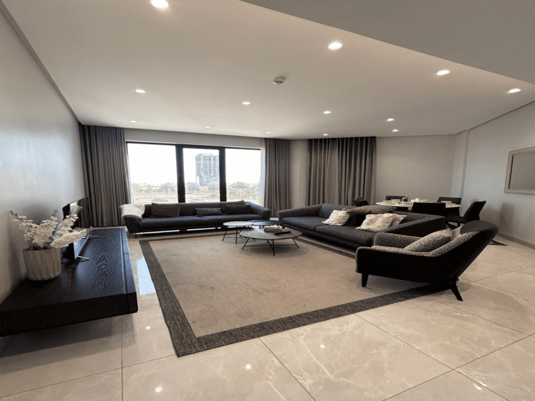 Luxury apartment with a modern living room featuring gray sofas, an armchair, a coffee table, and large windows with a city view. It has a neutral color palette and recessed lighting.