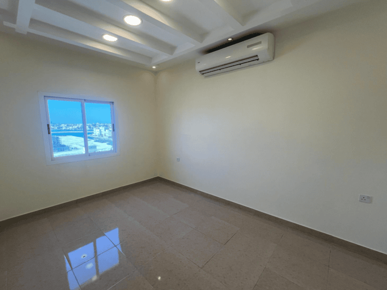 An empty room for rent in Hidd with tiled flooring, a window with a view of the city, and a wall-mounted air conditioner.