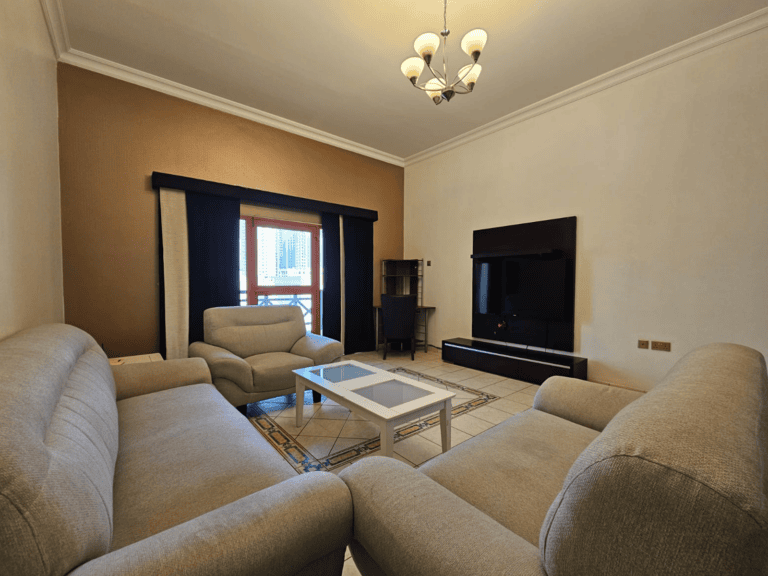 Modern living room available in a Juffair flat for rent, featuring beige walls, a three-seat sofa, two armchairs, a white coffee table, hanging chandelier, and a black TV