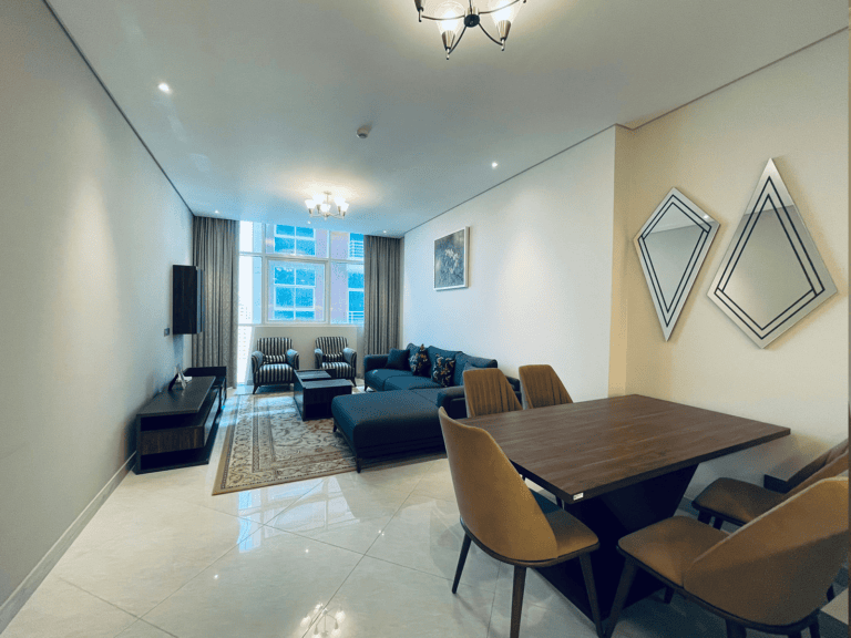 Modern living room in a luxury 1BR apartment in Juffair with a blue sofa, two chairs, a dining table, and a TV, featuring neutral decor and ample lighting.