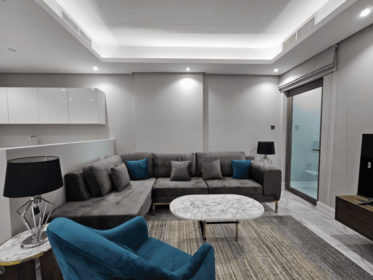 Modern living room in a Juffair flat for rent, featuring a large gray sectional sofa, a blue armchair, a marble coffee table, and black lamps, leading to a balcony.
