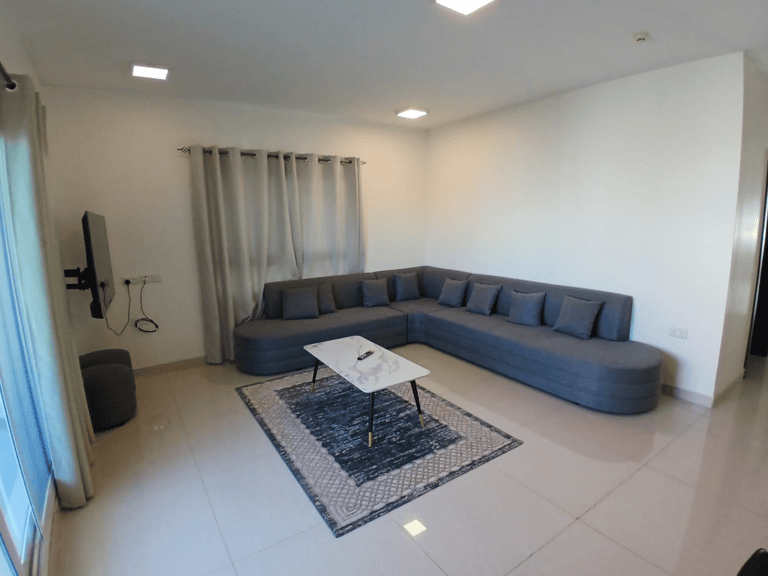 غرفة معيشة حديثة تحتوي على أريكة كبيرة مقطعية باللون الرمادي، وطاولة قهوة مستطيلة على سجادة زرقاء، وتلفزيون مثبت على الحائط على اليسار.