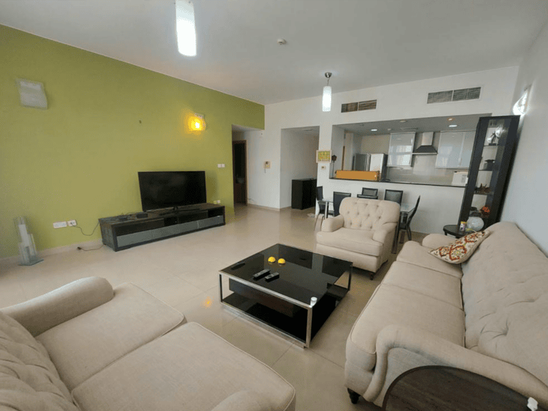 A modern, spacious living room in a flat in Amwaj with light furniture, a large TV, and an open kitchen in the background.