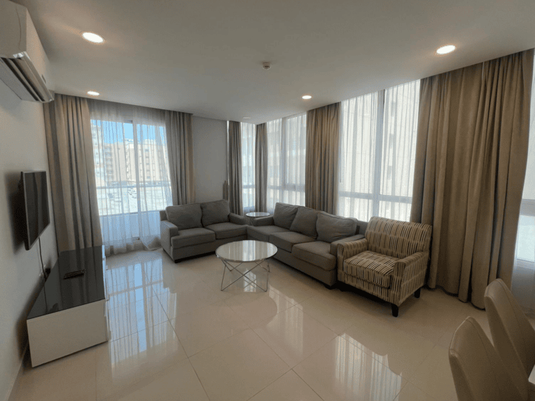 Modern living room in a 2 BHK apartment with a grey sectional sofa, striped chair, glass coffee table, and mounted tv, illuminated by luxury lighting from large windows with sheer curtains.