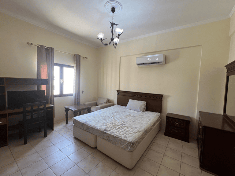 A simple bedroom in an apartment located in Busaiteen, with a double bed, two nightstands, a desk, a chair, and a wardrobe, featuring a ceiling fan and an air conditioning