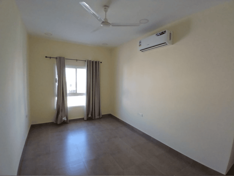 Flat for Rent: Empty room with beige walls, a window with curtains, ceiling fan, and an air conditioning unit in AL Hidd area.