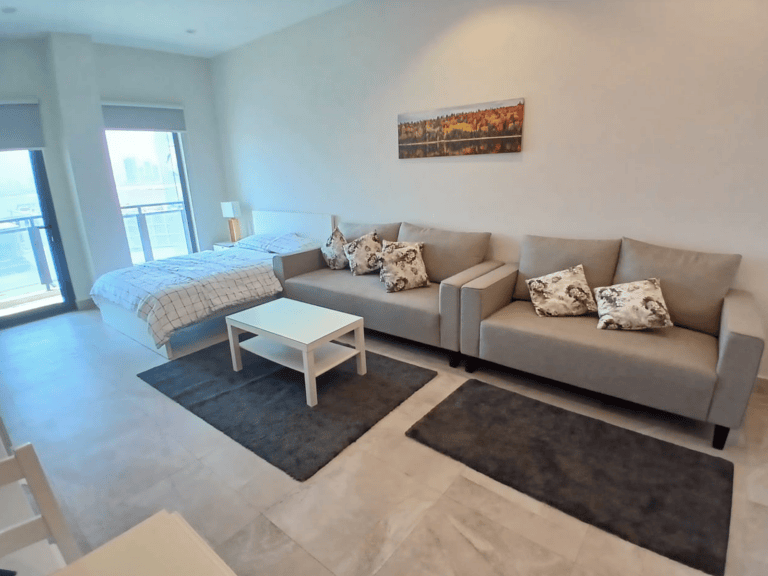 A luxury studio apartment in Busiteen with two gray sofas, a white coffee table, twin bed, wall art, and large windows providing natural light.
