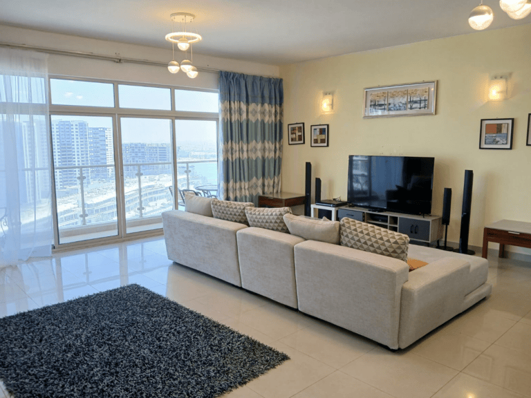 A modern flat in Amwaj with a large window overlooking a cityscape, featuring two beige sofas, a TV, and a blue rug.