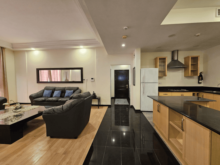 This modern flat for rent in Juffair boasts an open-plan living area with wooden flooring, a black sofa set, and a sleek black-tiled kitchen. The kitchen features wooden cabinets, a fridge, and a range hood. A hallway and door are visible in the background.