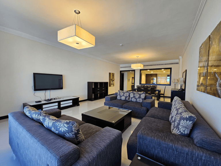 A spacious living room in a Juffair flat for rent, featuring blue sofas, a coffee table, and a wall-mounted TV. The room is well-lit with large overhead light fixtures and leads into a dining area with a view of the kitchen.