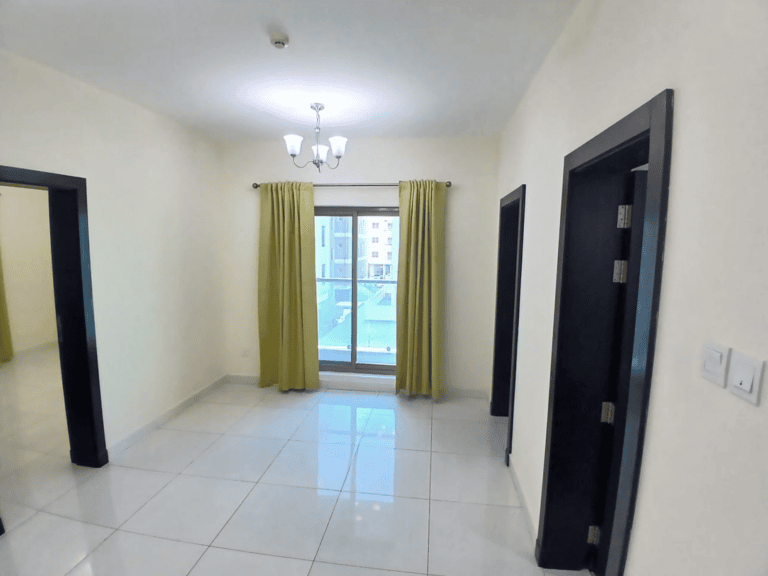A minimally furnished flat in New Hidd, featuring tiled flooring, a window with yellow curtains, and a ceiling light fixture. The room is in very good condition and includes two doorways leading to adjacent spaces.