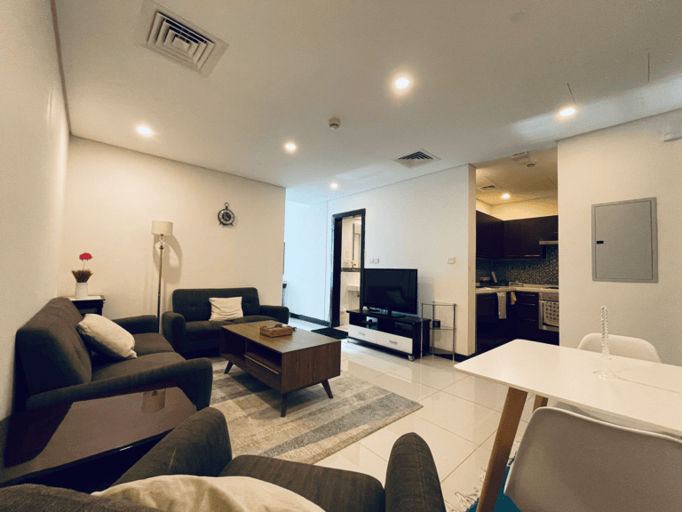In a luxury 1BR apartment in Juffair, a modern living room boasts a grey sofa set, wooden coffee table, floor lamp, and wall-mounted TV. This stylish space flows seamlessly into an open kitchen and dining area featuring white chairs and a sleek table.