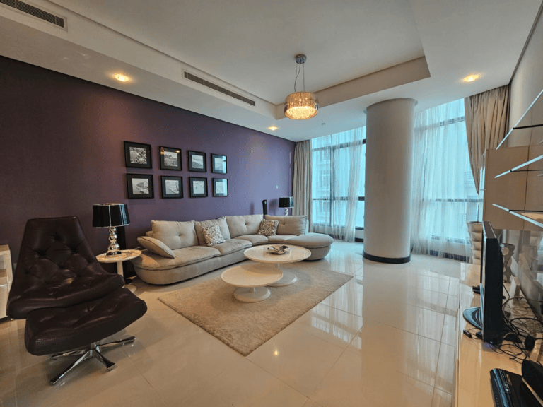 A modern living room in a Seef flat for rent, featuring a sectional sofa, lounge chair, coffee tables, and a TV. The room boasts a purple accent wall adorned with framed art and a large window allowing natural light to flood the space.