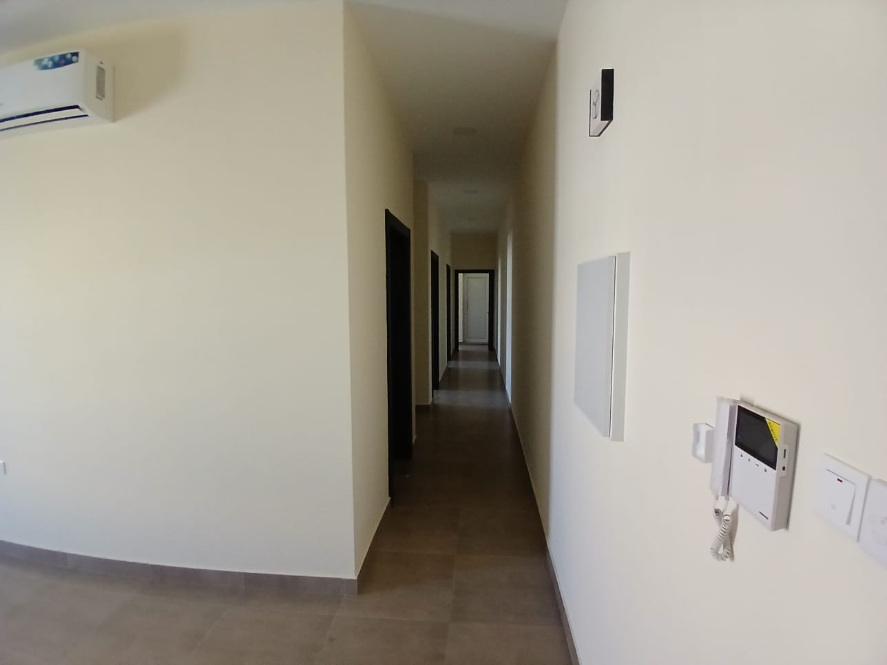 A long, narrow hallway in a modern building with beige walls, tiled floor, and doors on the left side in the AL Hidd area. An air conditioning unit and intercom system are visible.