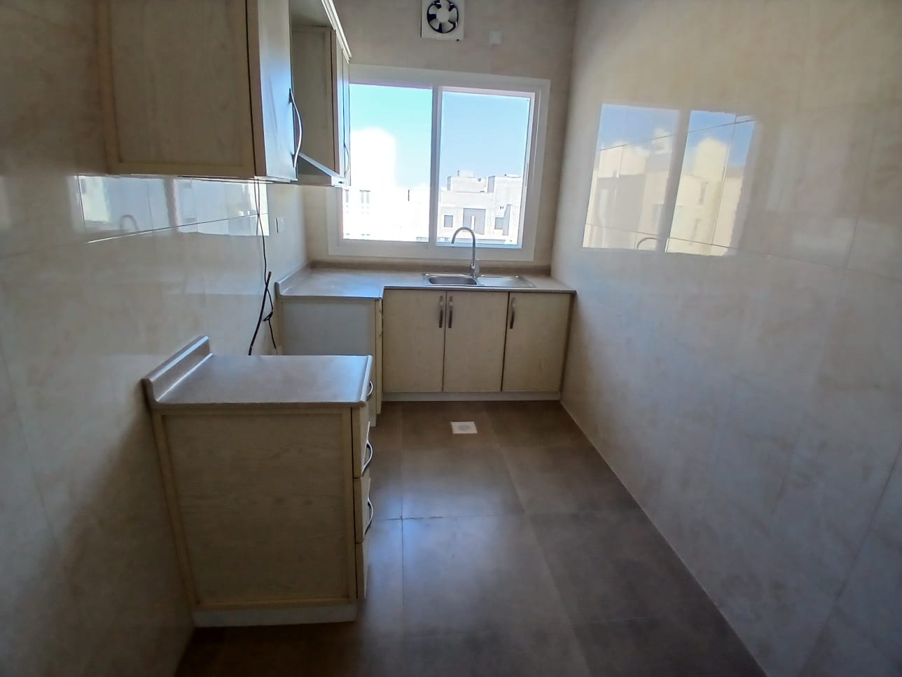 A small, empty kitchen with beige tiles in AL Hidd area, featuring a window, built-in cabinets, and a detached countertop on the left.