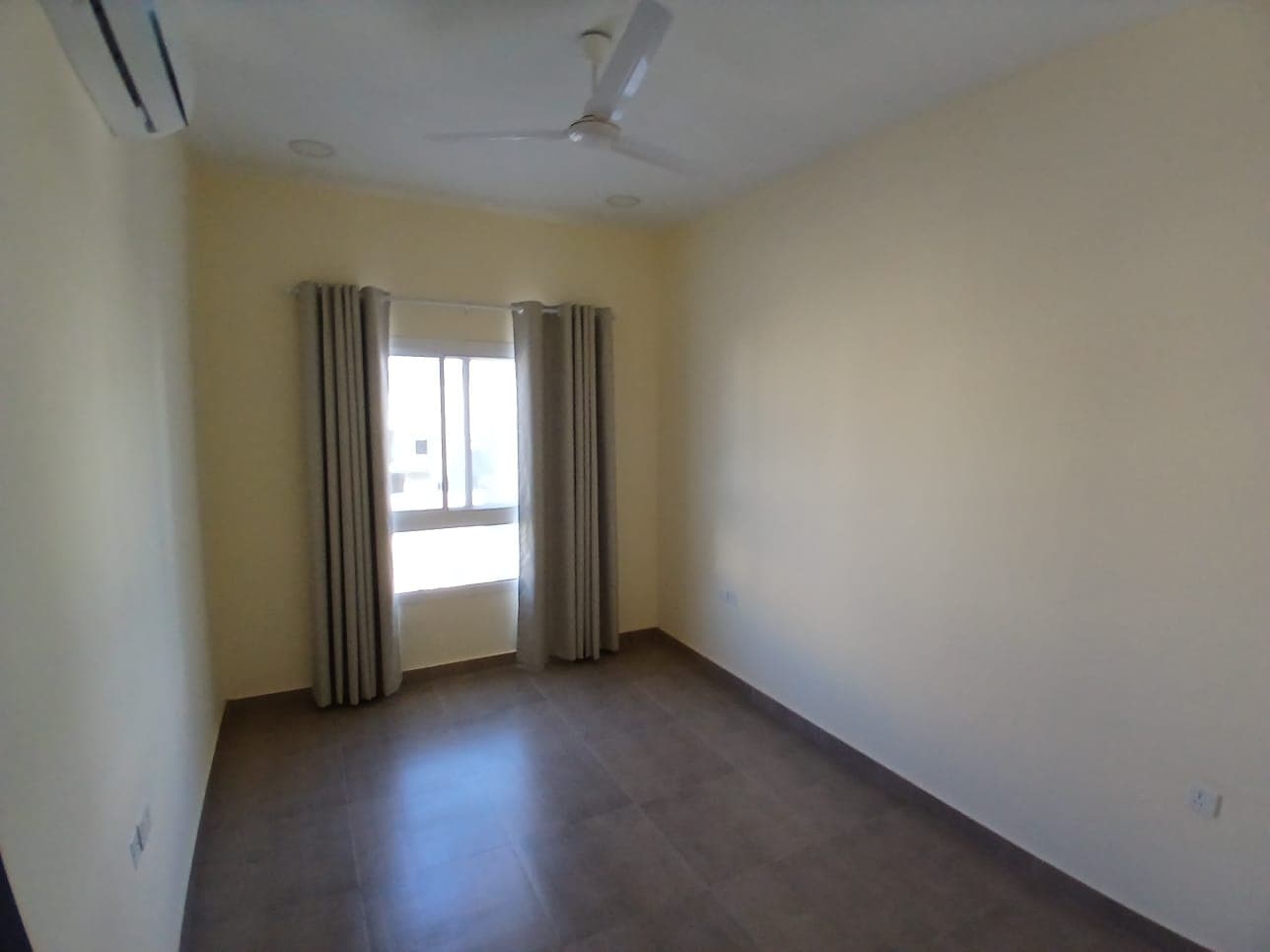 Empty flat for rent in AL Hidd with beige walls, a tiled floor, a ceiling fan, and a window with curtains.