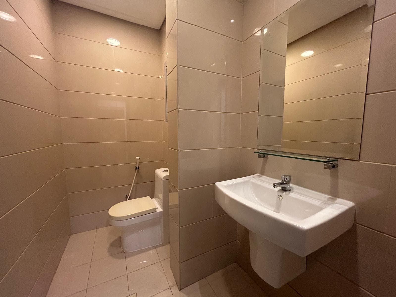 A small bathroom in a 3 BDR Apartment with beige tiles, featuring a toilet, a wall-mounted sink with a glass shelf, and a mirror.