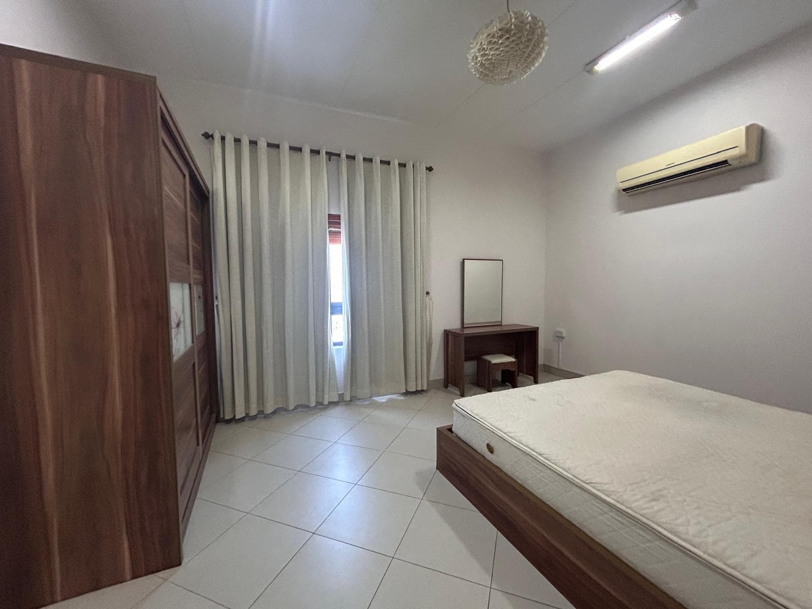 A minimalistic bedroom in a luxury apartment featuring a large bed, an air conditioner, wooden wardrobe, a small desk with a mirror, tiled flooring, and a window with white curtains.