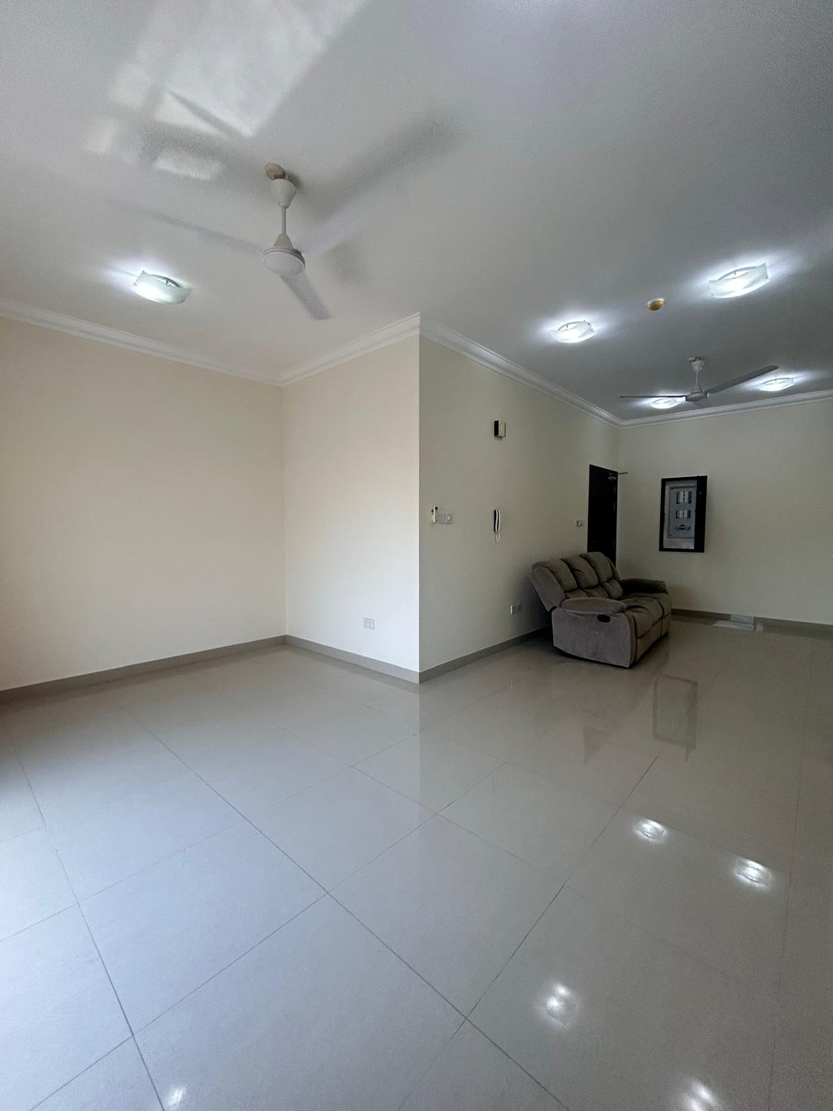 Spacious, empty room in a 3 BDR Apartment with glossy tiled floor, beige walls, and a gray sofa near the entrance.