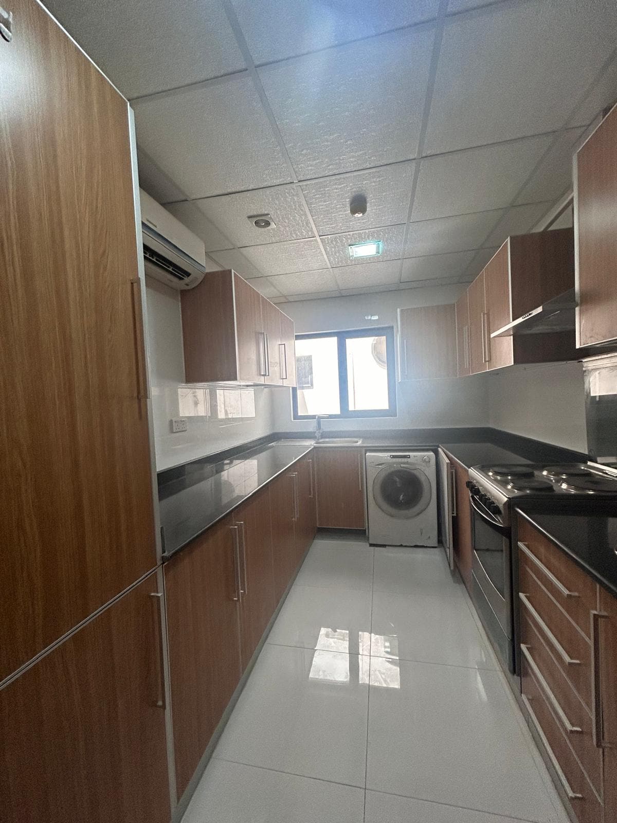 A modern kitchen in a 3 BDR apartment for rent, with wooden cabinets, stainless steel appliances, including a washing machine, and a window at the end.