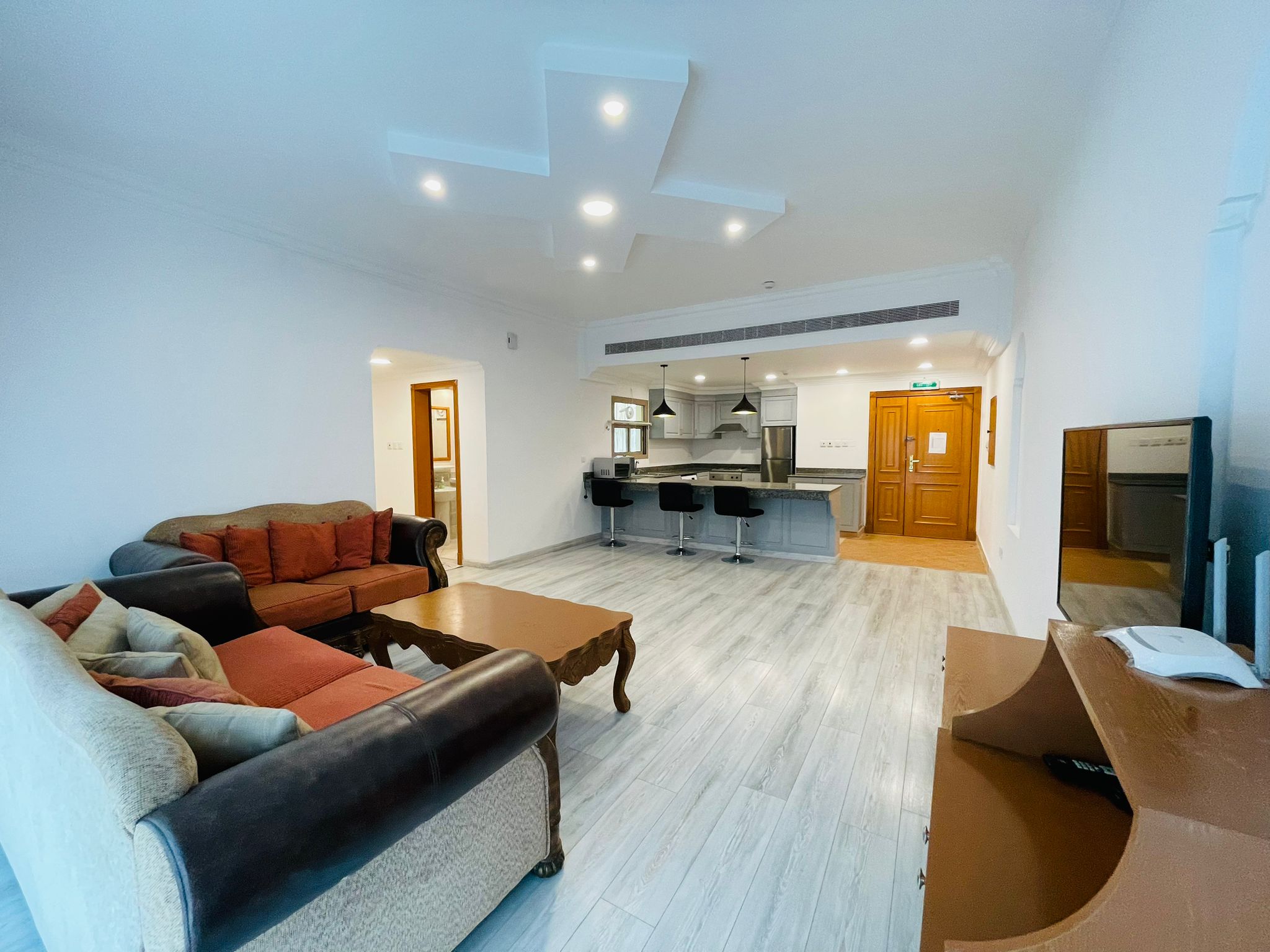 Spacious living room with two brown sofas, a coffee table, and an open kitchen with bar stools in a furnished apartment. The room features modern lighting and wooden flooring.