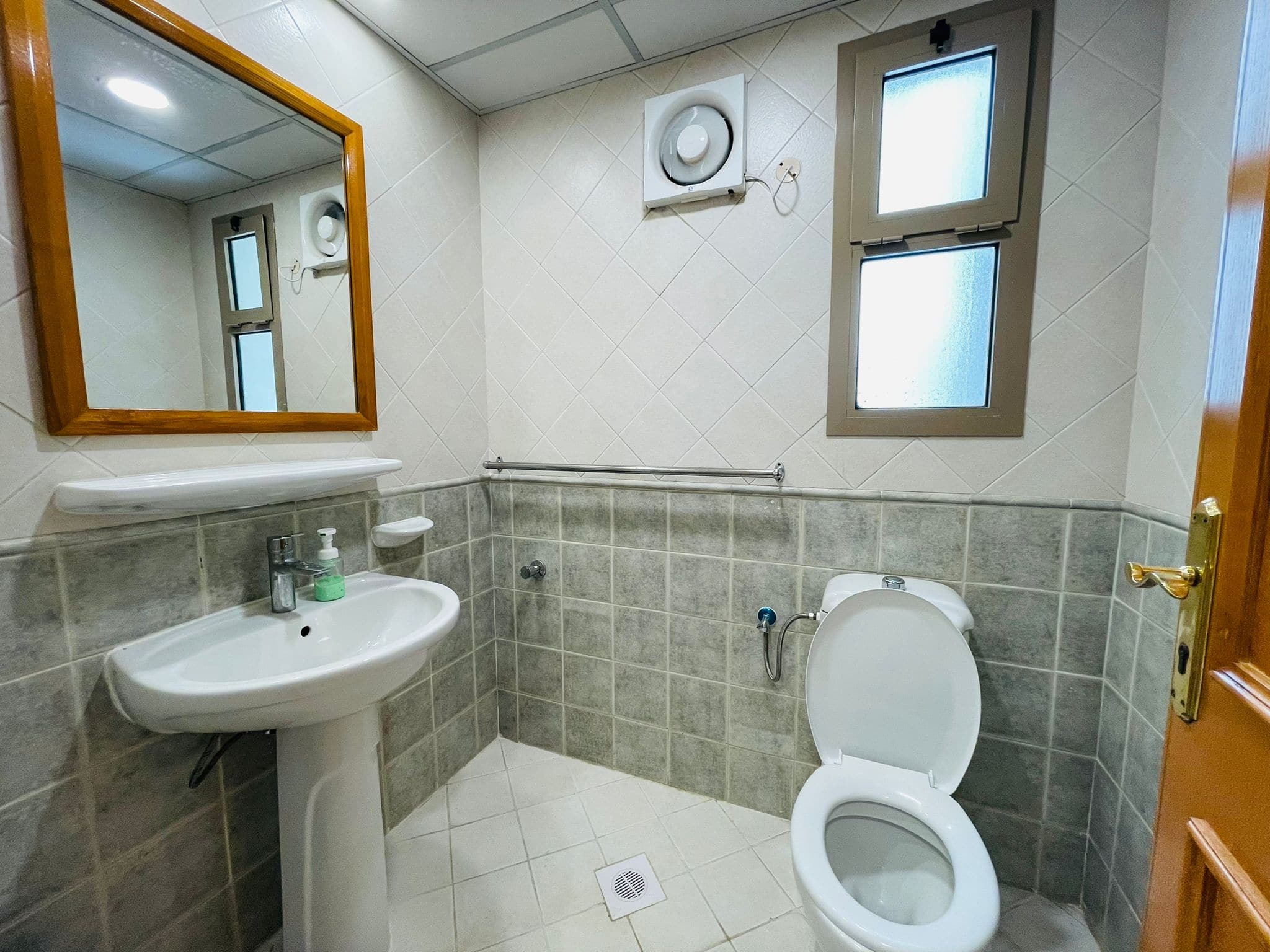 A clean, modern bathroom in a furnished apartment with tiled walls featuring a toilet, sink, mirrored medicine cabinet, and frosted windows.