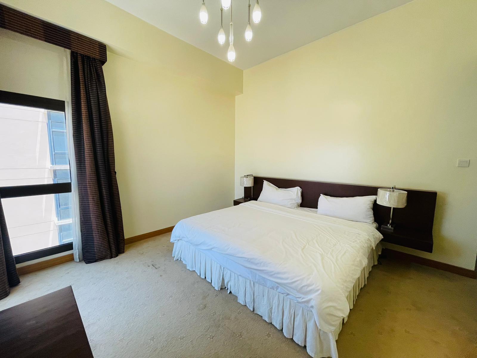 A stylish furnished apartment in the Sanabis area featuring a modern bedroom with a large bed adorned by white bedding, two nightstands with lamps, a window, and a chandelier.