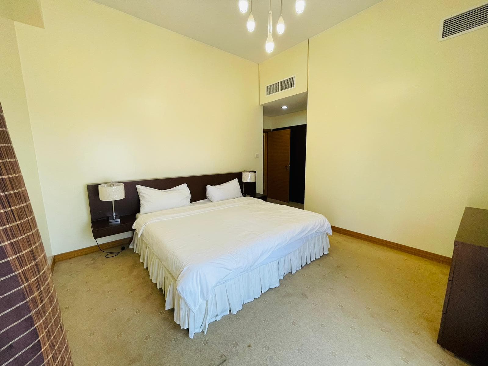 Modern minimalist bedroom in a furnished apartment with a large double bed, white bedding, two bedside lamps, and creamy yellow walls.