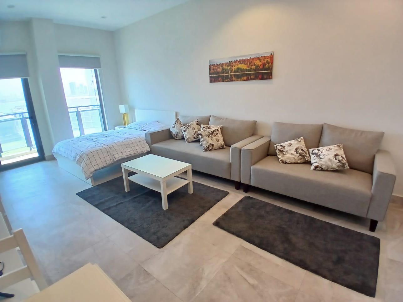 Modern luxury studio apartment interior for rent in Busiteen, featuring a beige sofa, a coffee table, black rugs, and a bed by large windows with natural light.