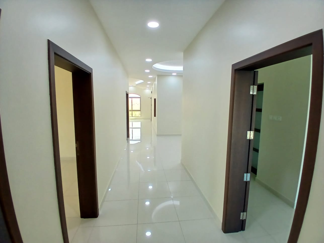 A brightly lit hallway in a brand new apartment with glossy white tiles, featuring several wooden doors and a large mirror on the right wall.