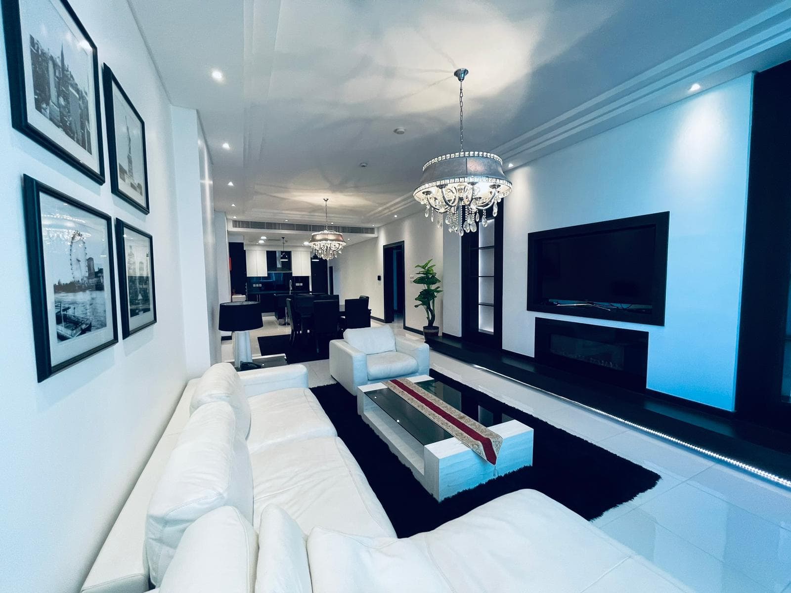 Elegant living room in a luxury apartment with white sofas, a chandelier, and framed pictures on the walls, extending into a dining area.