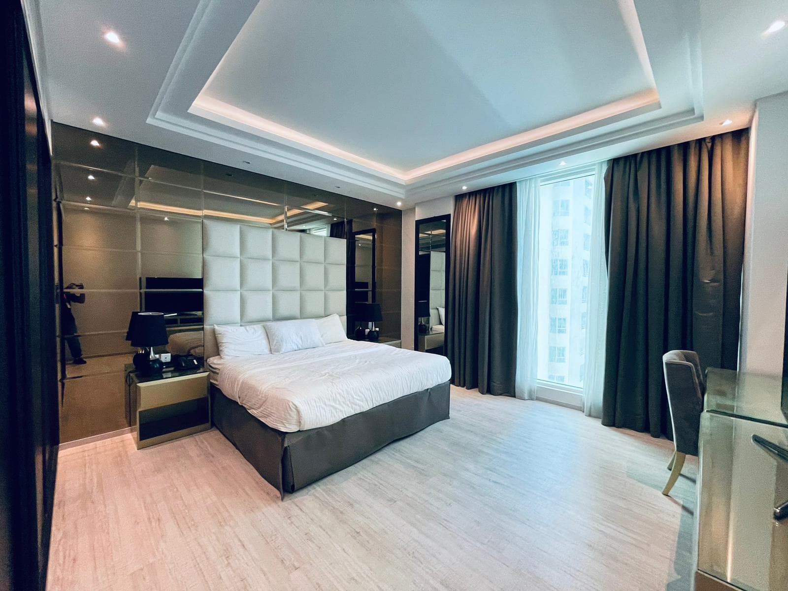 Luxury bedroom with a large bed, plush headboard, dark curtains, and a desk area; highlighted by elegant, soft lighting and reflective surfaces.